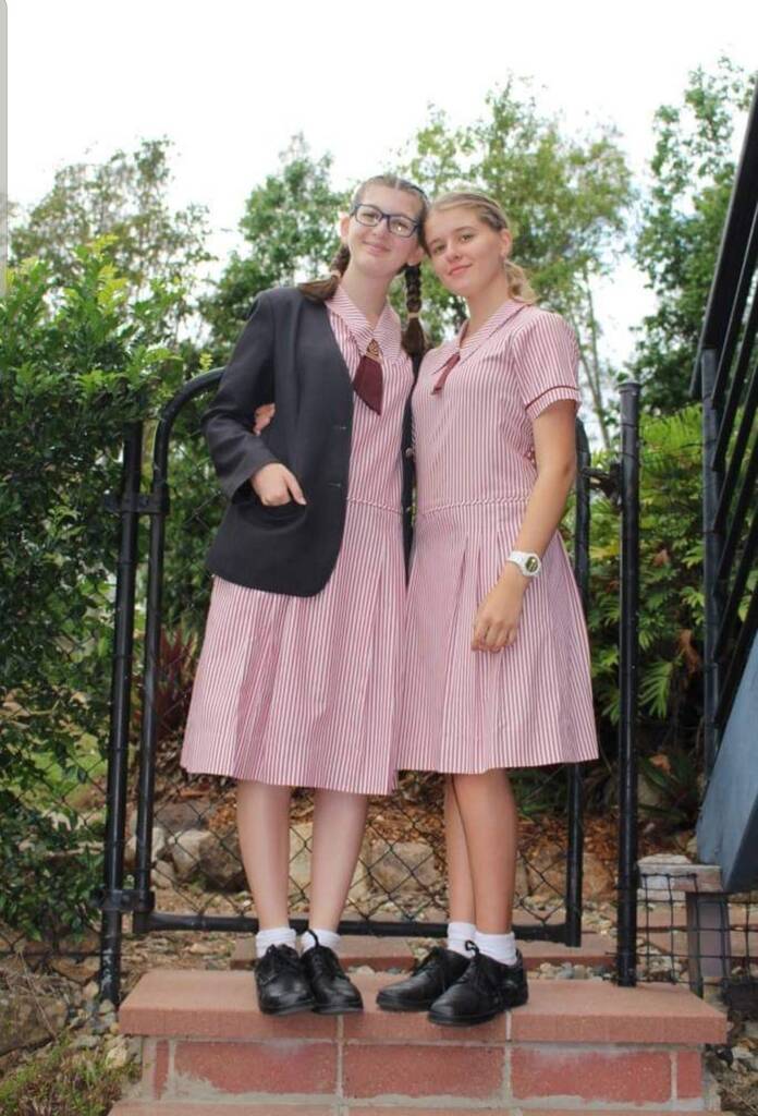Eva Hopewell at school, standing with a classmate in matching pink-and-white striped school uniforms near a school gate with greenery in the background