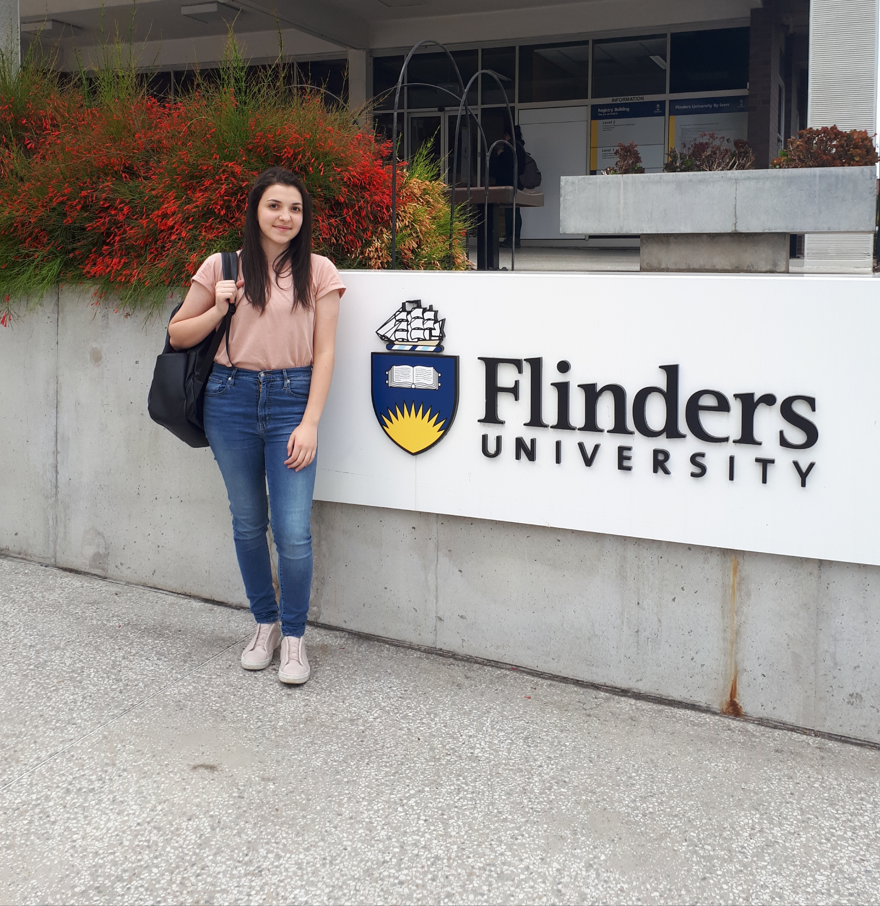 Alissar Azar standing beside the Flinders University sign on campus grounds with buildings and greenery in the background