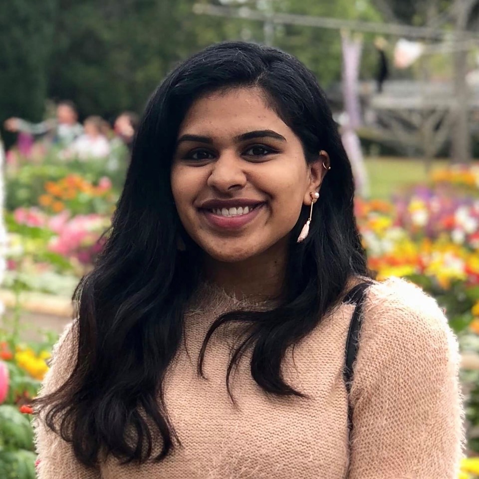 Angel Maliakkal standing in a garden setting, wearing a soft pink knit top with colourful flowers and greenery in the background