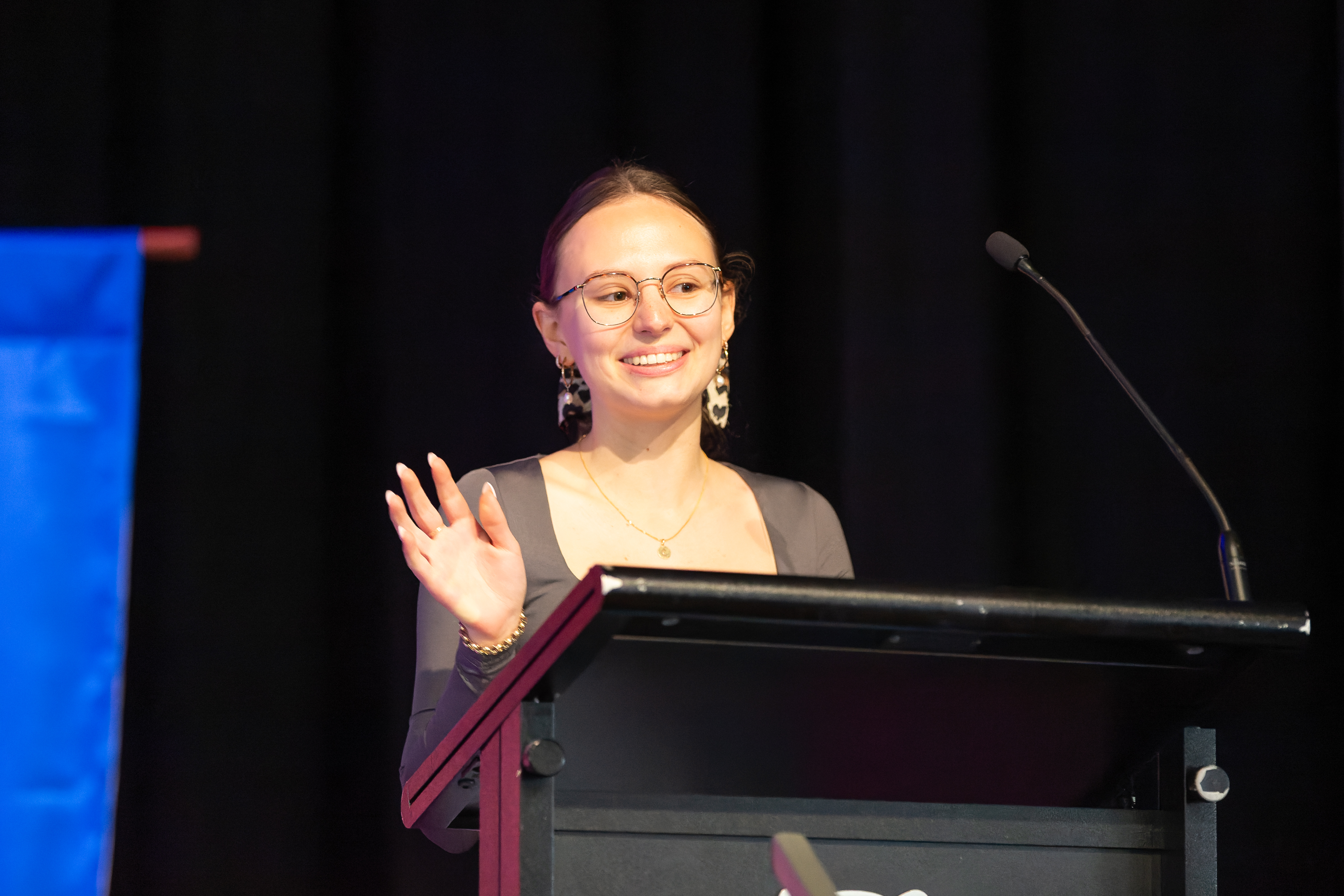 Caitlyn Douglas speaking at a podium on a stage with a microphone and banner in the background