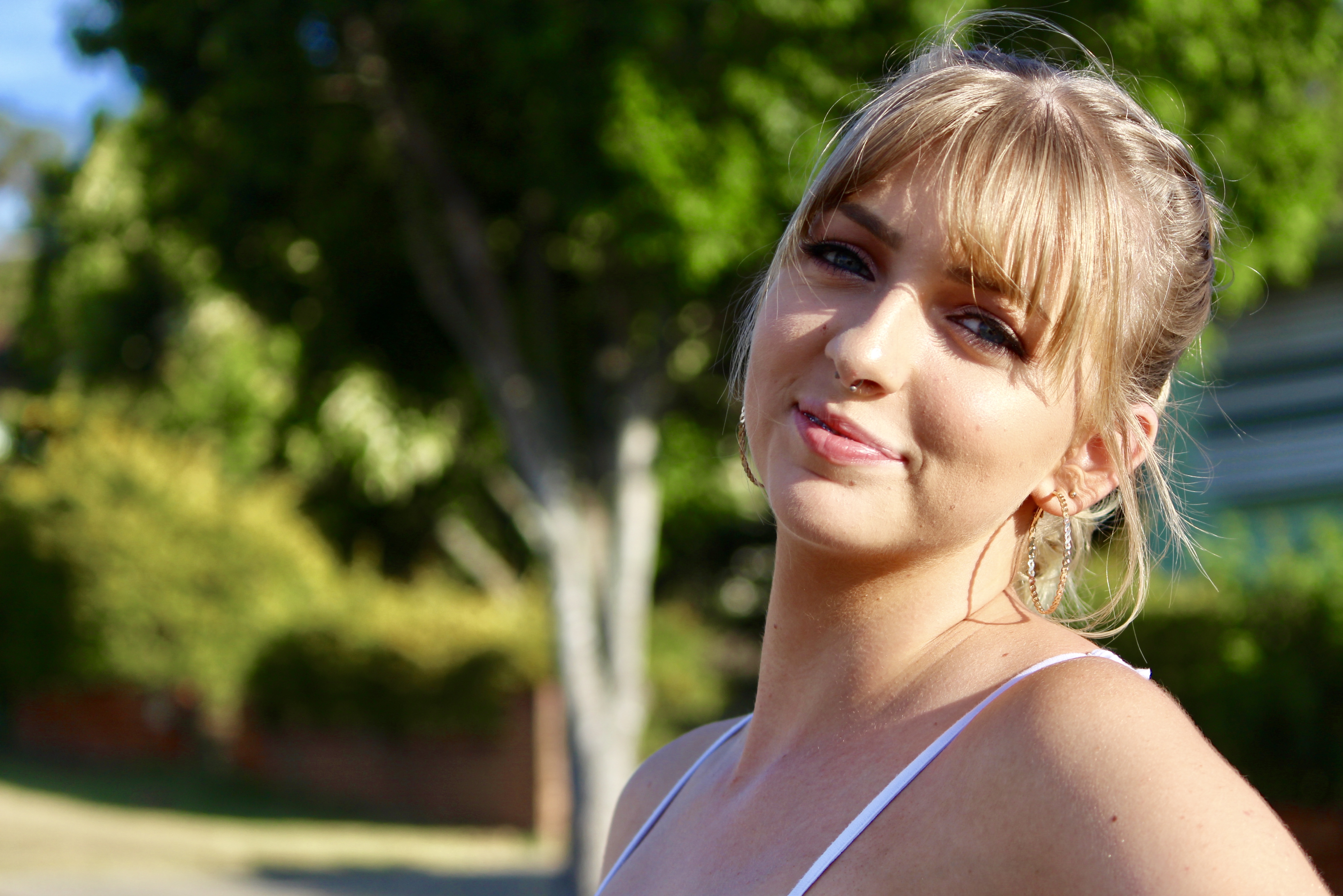 Morgan standing outdoors in soft sunlight, wearing a light sleeveless top with greenery and a residential street in the background