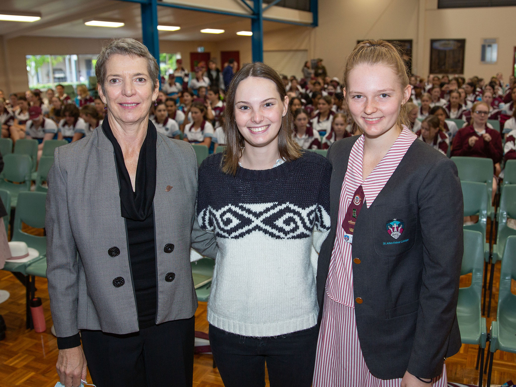 SJFC alumni Maya Rees, College Captain of the Class of 2018, standing with staff members in a school hall during an assembly with students seated in the background
